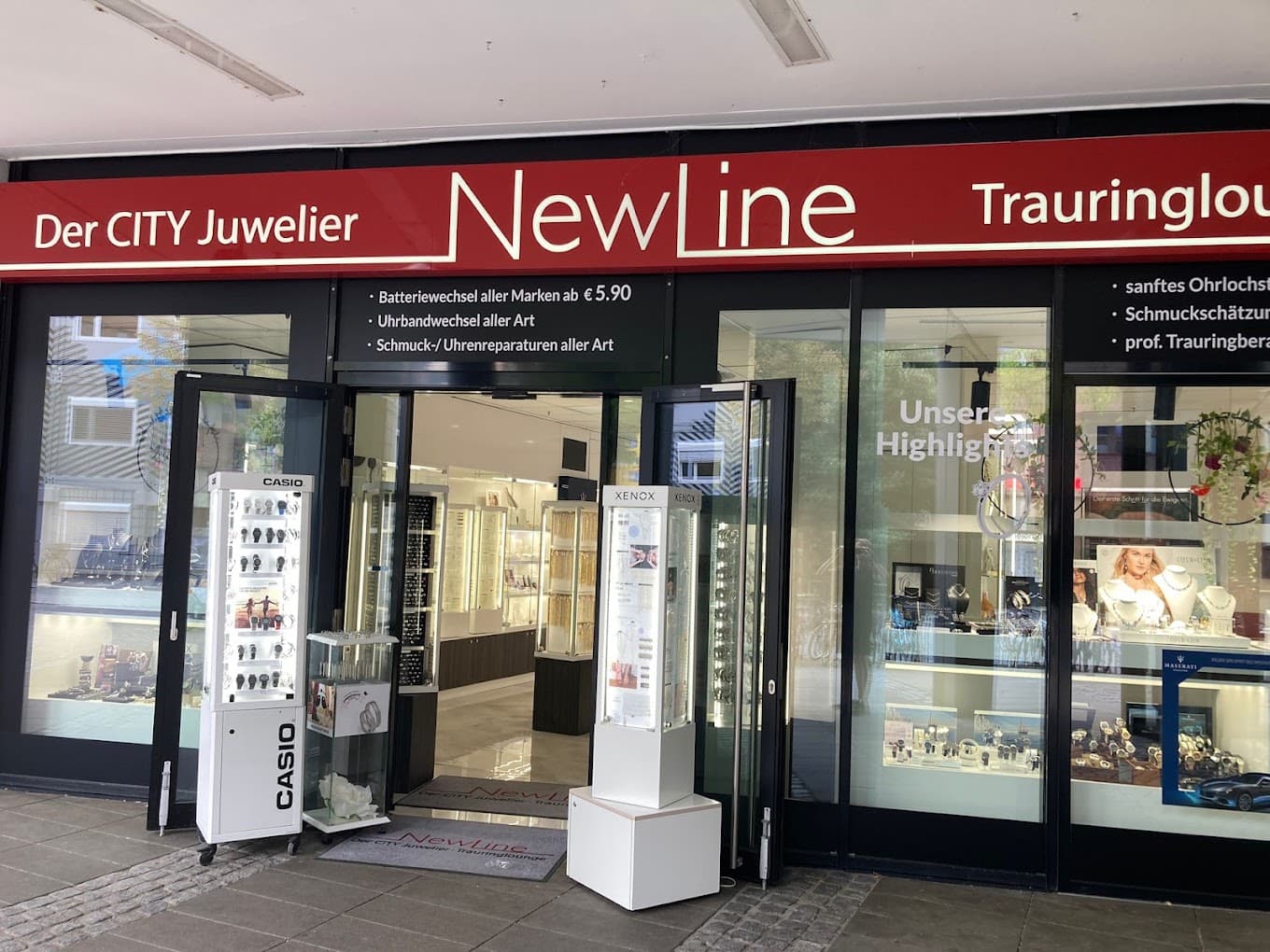Ladenfront des Juweliers NewLine mit roter Schrifttafel, geöffneter Eingangstür, Casio-Uhrenständern und Schaufenstern mit Schmuck und Uhren